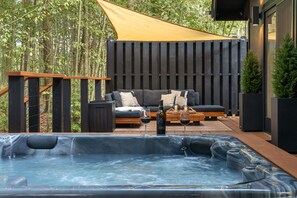 A hot tub with a wooden deck, a sofa with cushions, and a wooden fence in a wooded area.