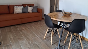 A modern living room with a sofa, a wooden dining table with chairs, a patterned rug, and a wall with a 'Welcome' sign and a 'Ceci n'est pas un hotel' sign.