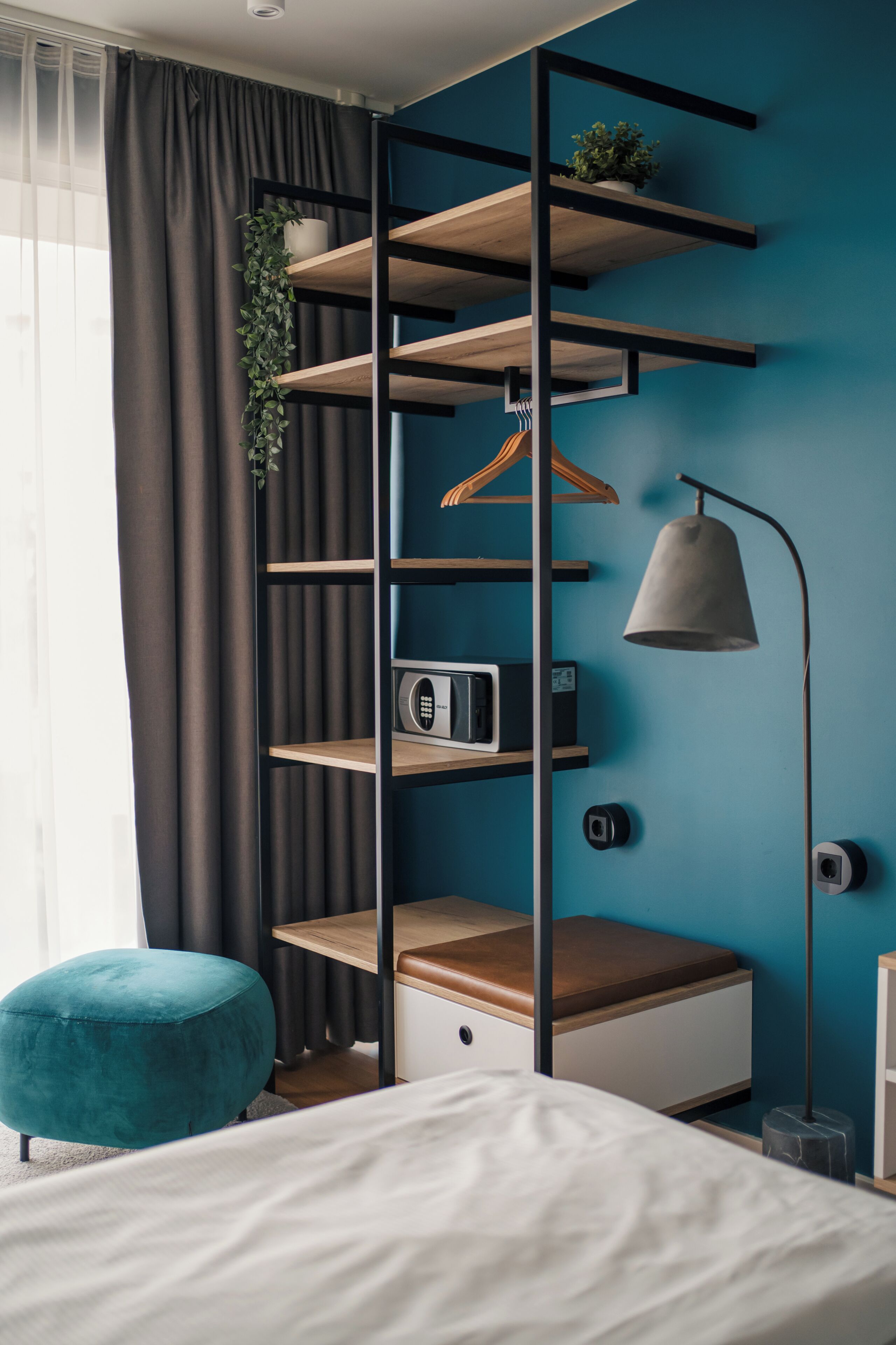 Un dormitorio con una cama, un reposapiés color turquesa, un banco de madera, un estante en la pared con una planta, una lámpara colgante y un radio.