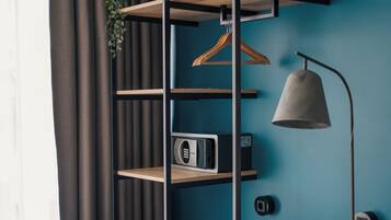 Un dormitorio con una cama, un reposapiés color turquesa, un banco de madera, un estante en la pared con una planta, una lámpara colgante y un radio.