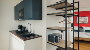 Una kitchenette compacta con microondas, lavamanos y cafetera, una cama frente a una pared roja y un estante con una planta colgante.