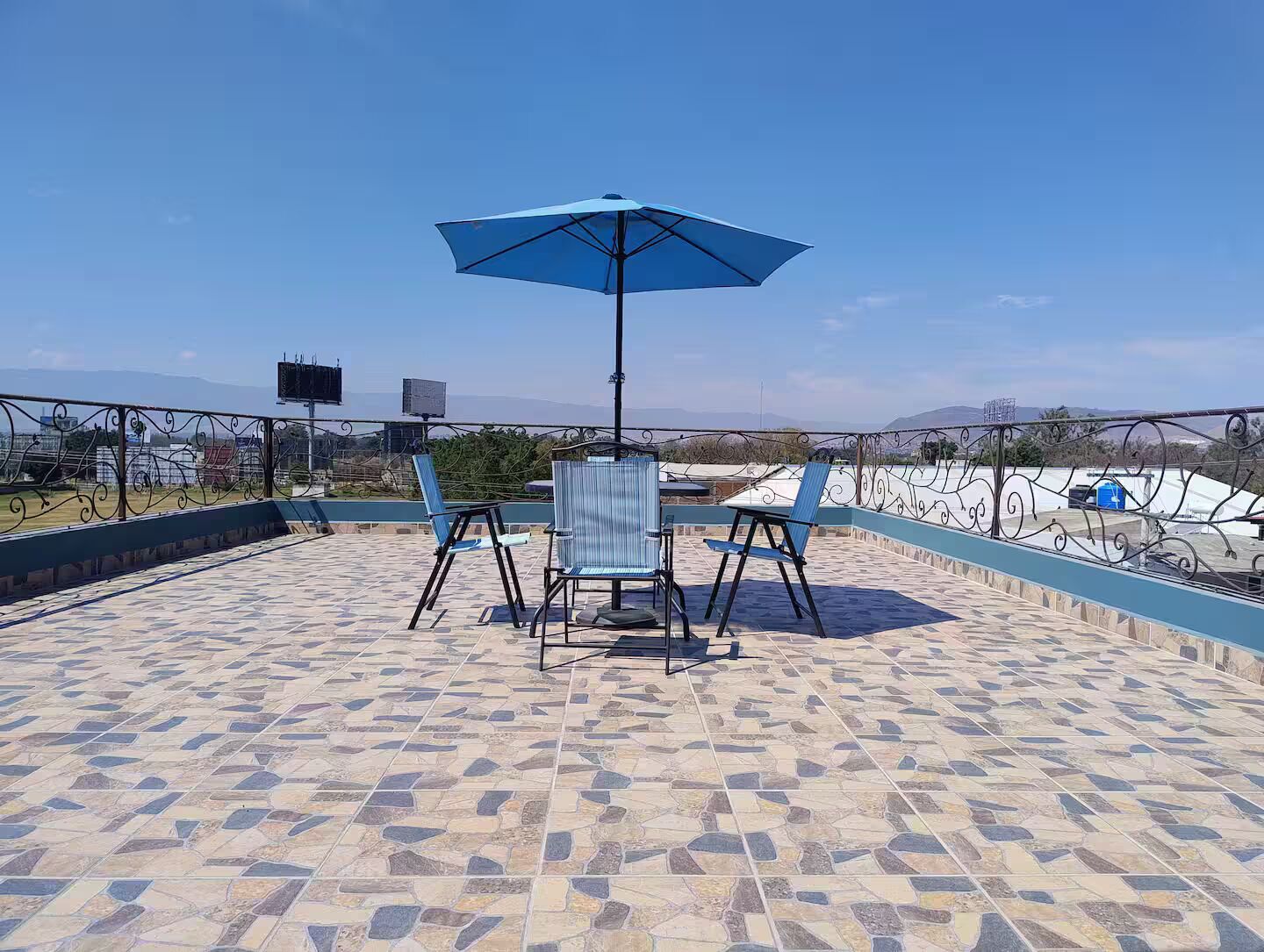An outdoor patio with a table and chairs, a blue umbrella, and a view of the landscape.