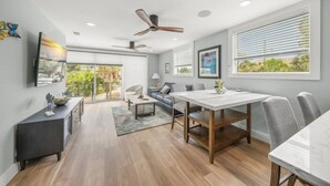A modern living room with a dining area, large windows, ceiling fan, and artwork on the walls.