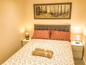 Ein Bett mit blumigem Überwurf, einem Kopfteil, zwei Nachttischen mit Lampen und einem Waldgemälde im Rahmen darüber.
