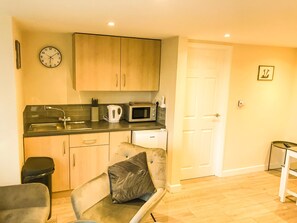 A compact kitchen with a sink, microwave, kettle, and cabinets. There is a clock on the wall and a door to the right. - Apartment Sleeps 4 With Parking (Blakeney)