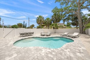 A swimming pool with lounge chairs and a table, surrounded by a white fence and palm trees.