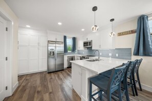 A modern kitchen with white cabinetry, a stainless steel refrigerator, a microwave, and a sink. There are blue chairs and a bar counter.