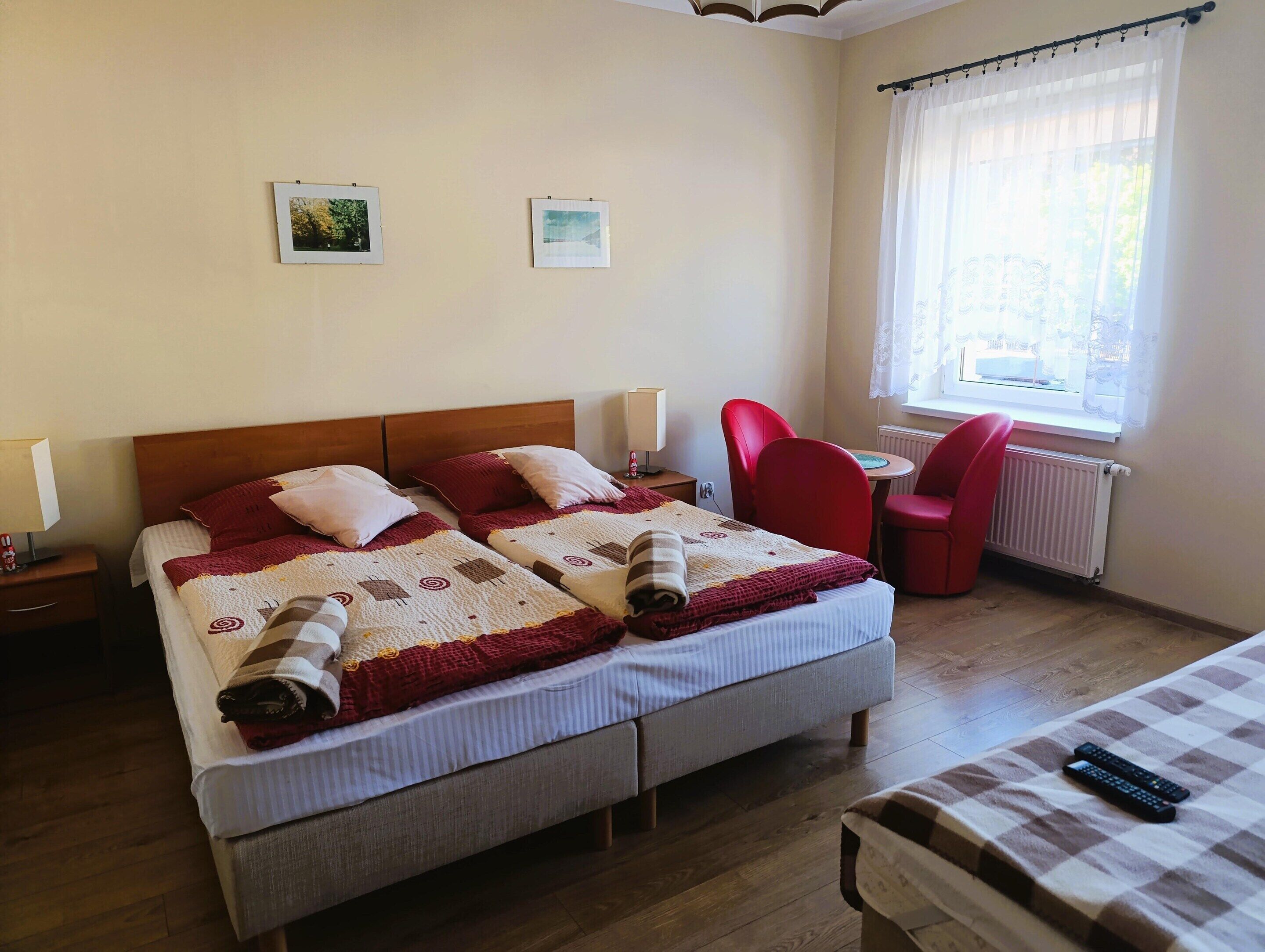 A double bed with a quilt, two bedside tables with lamps, a red chair, a small table with a chair, and a window with curtains.