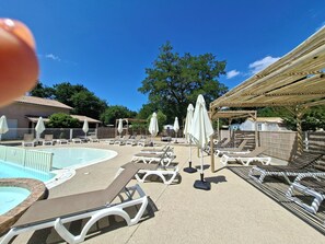 Et poolområde med liggestole og parasoller, omgivet af en træpergola og træer.