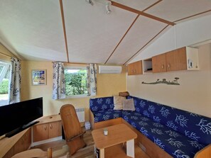 A compact living space with a sofa, a television, a wooden cabinet, and a window with curtains. - Mobile home rental 4 pers Carcans (Carcans)