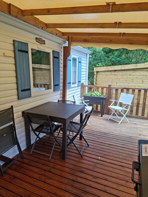 A covered deck with a table and chairs, a wooden fence, and a house with blue shutters. - Mobile home rental 4 pers Carcans (Carcans)