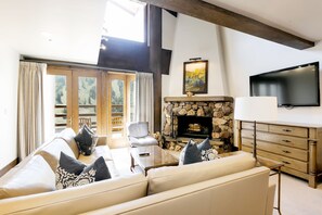 Una sala de estar con chimenea de piedra, un conjunto de sofás, un televisor y un ventanal con cortinas.