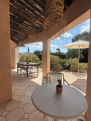 Eine überdachte Terrasse mit Tisch und Stühlen, einem hängenden Korb und Blick auf den umliegenden Garten.