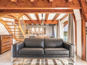 A modern interior with a leather sofa, wooden beams, a kitchen, and a staircase. - Duplex 90m² | Alsatian courtyard & parking | Route des Vins (Marlenheim)