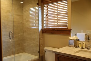 A bathroom with a glass-enclosed shower, a toilet, a sink with a marble countertop, and a window with blinds. - Upgraded Deluxe Mountain Studio (Park City)