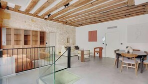 Une pièce spacieuse avec un plafond haut, des poutres apparentes, un escalier en verre, un coin repas avec une table et des chaises, et un mur blanc agrémenté d’une porte simple et d’un petit meuble.