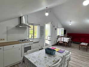 Cozinha compacta com fogão a gás, exaustor e janela. Há uma área de jantar com mesa e cadeiras, e um sofá vermelho.