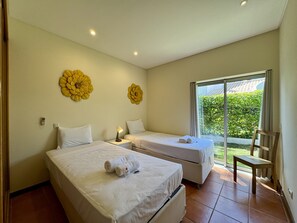 Ein Hotelzimmer mit zwei Betten, einem kleinen Tisch, einem Sessel und einem großen Fenster mit Blick auf Grünflächen.