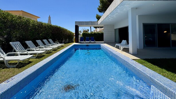 A modern pool area with lounge chairs, a clear blue pool, and a building with large windows. - Albufeira Balaia Villa 2 by Homing (Albufeira)