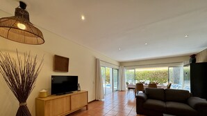 A spacious living room with a flat-screen TV on a wooden cabinet, a dark sofa, a coffee table, and a large window offering a view of the outdoors. - Albufeira Balaia Villa 2 by Homing (Albufeira)