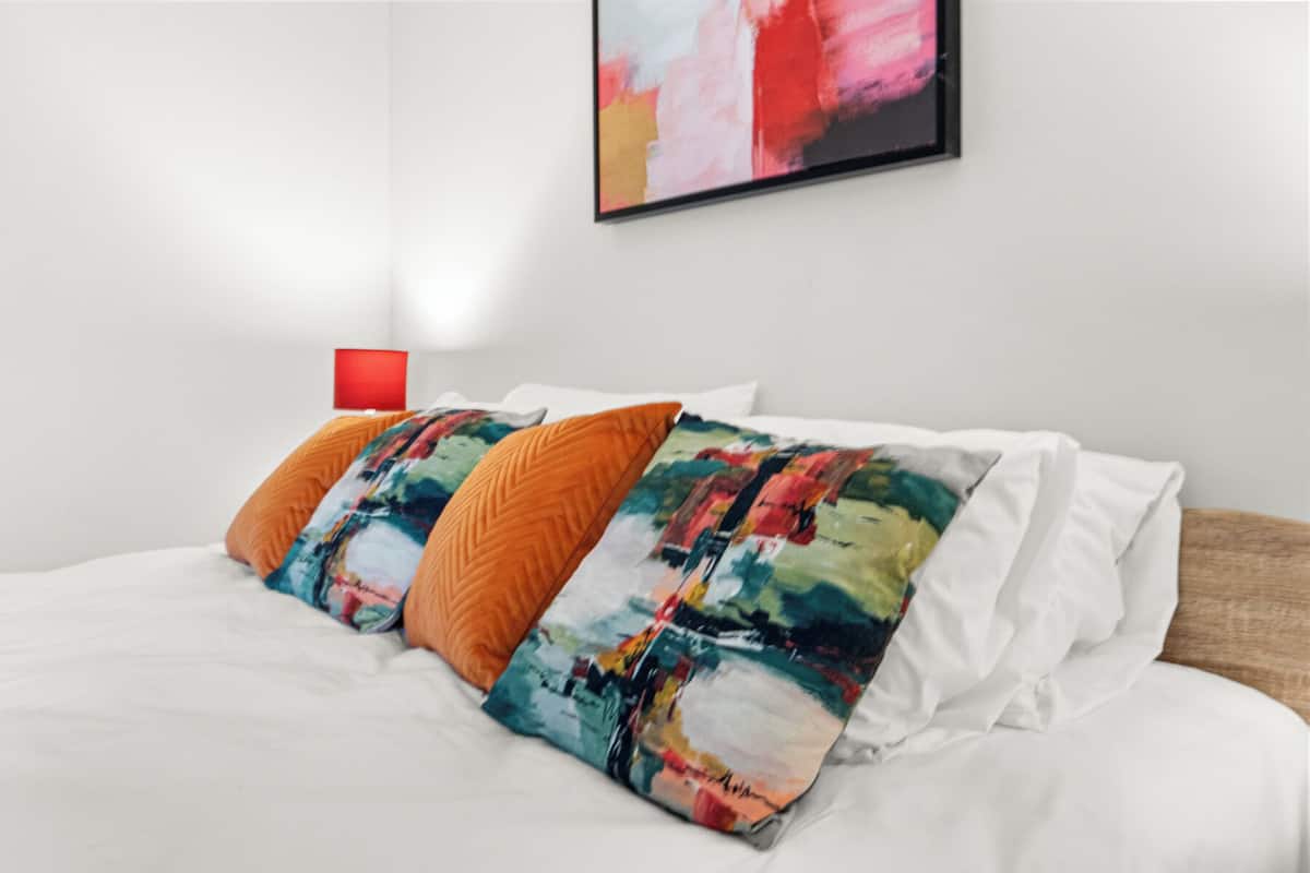 Un lit avec une literie blanche, des coussins colorés et une lampe rouge sur un mur blanc.