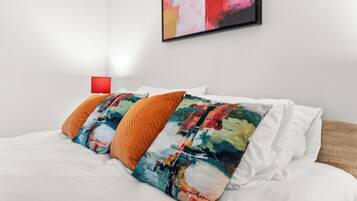 A bed with white bedding, colorful pillows, and a red lamp on a white wall.