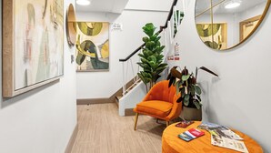 Un pasillo con una escalera, un espejo grande, una planta, un sillón naranja y un otoman redondo con revistas.
