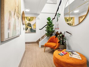 Un pasillo con una escalera, un espejo grande, una planta, un sillón naranja y un otoman redondo con revistas.