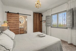A bedroom with a bed, a bedside table, a mirror, a chandelier, a window with curtains, and a view of the outdoors. - The View – Villa with Private Pool, Walking Distance to Cómpeta (Cómpeta)