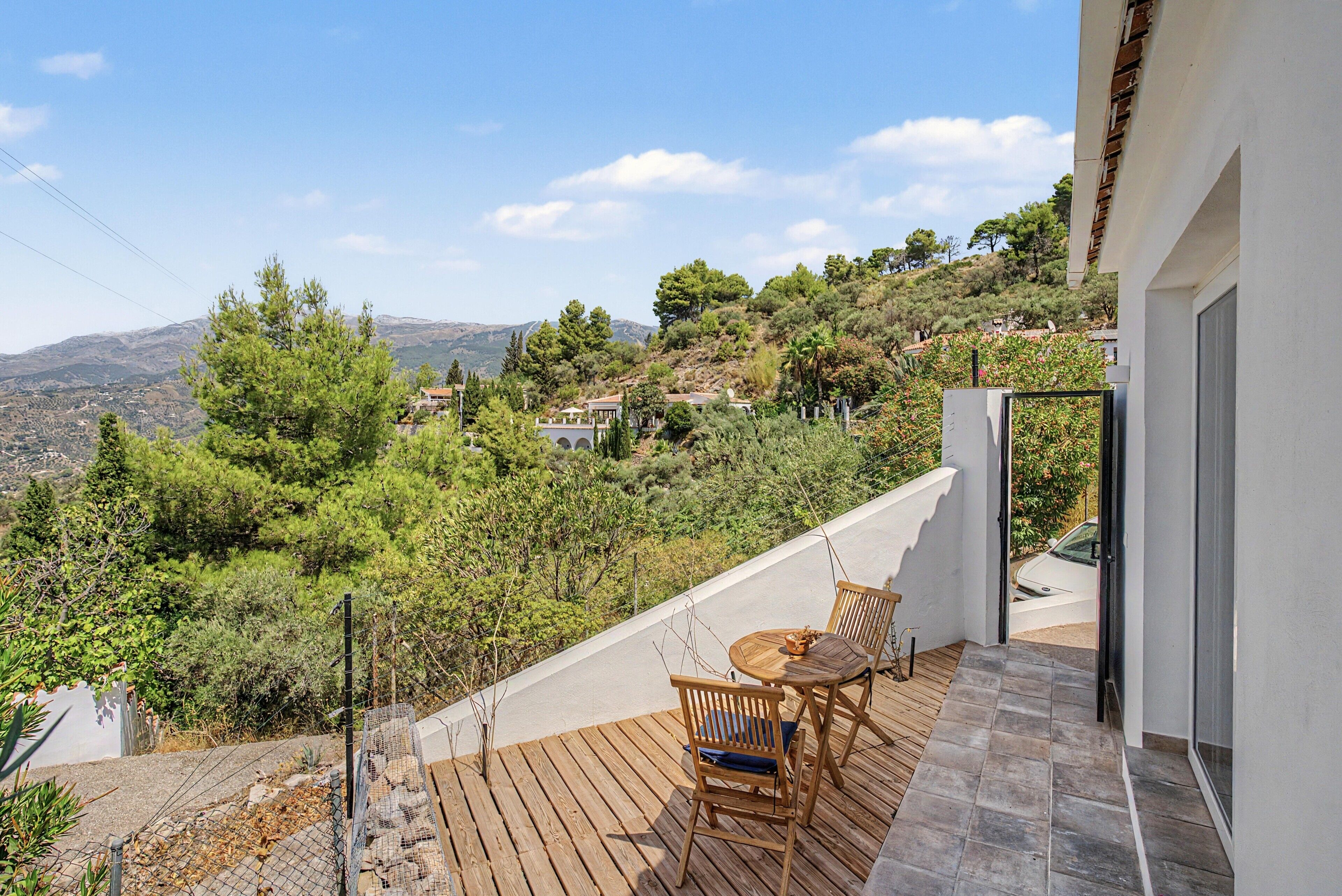 Un balcón con una mesa y sillas de madera, con vistas a una colina con árboles y casas.