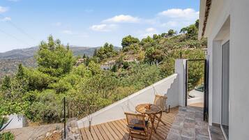 Un balcón con una mesa y sillas de madera, con vistas a una colina con árboles y casas.