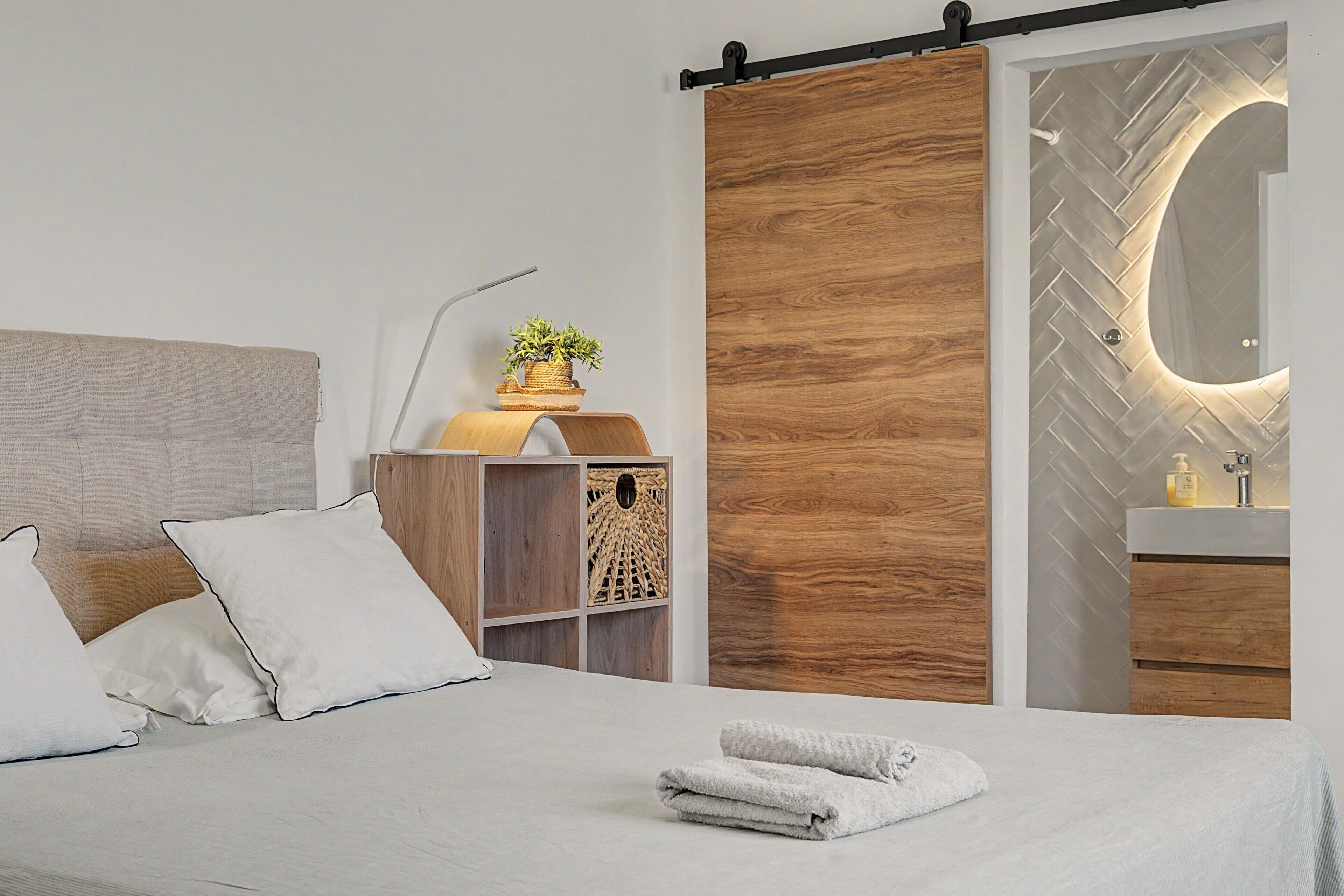 Un dormitorio moderno con cama, una mesita de noche con una planta, un panel de madera corredero y un baño con salpicadero de azulejos tipo espiga.