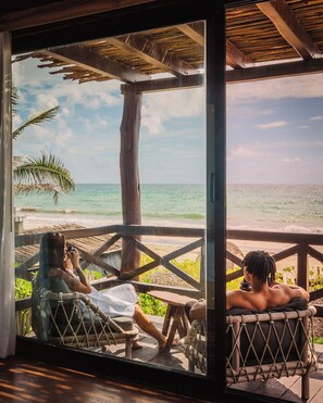A wooden deck with a view of the ocean, a palm tree, and a person sitting on a hammock. - Oceanfront Getaways Start Here Pool, Jacuzzi & Gym (Tulum)