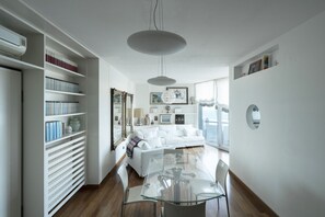 A modern living room with a glass dining table, white sofas, and a bookshelf.