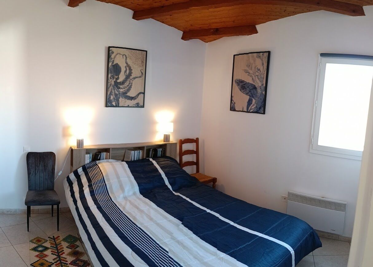 A bedroom with a bed, a chair, two wall-mounted lamps, and two framed artworks.