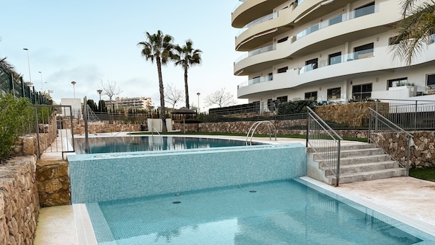 A swimming pool with a stone wall and steps leading to a building with balconies. - Apartment 'Bahia' with Shared Pool, Wi-Fi and Air Conditioning (Els Arenals del Sol)