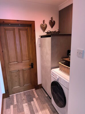A laundry room with a washing machine, wooden door, and decorative heart wall art. - Charming 2-bedroom cottage in Inverness perfect for relaxing stays (Inverness)