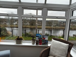 A conservatory with a view of a river, potted plants, and travel brochures on the windowsill. - Charming 2-bedroom cottage in Inverness perfect for relaxing stays (Inverness)