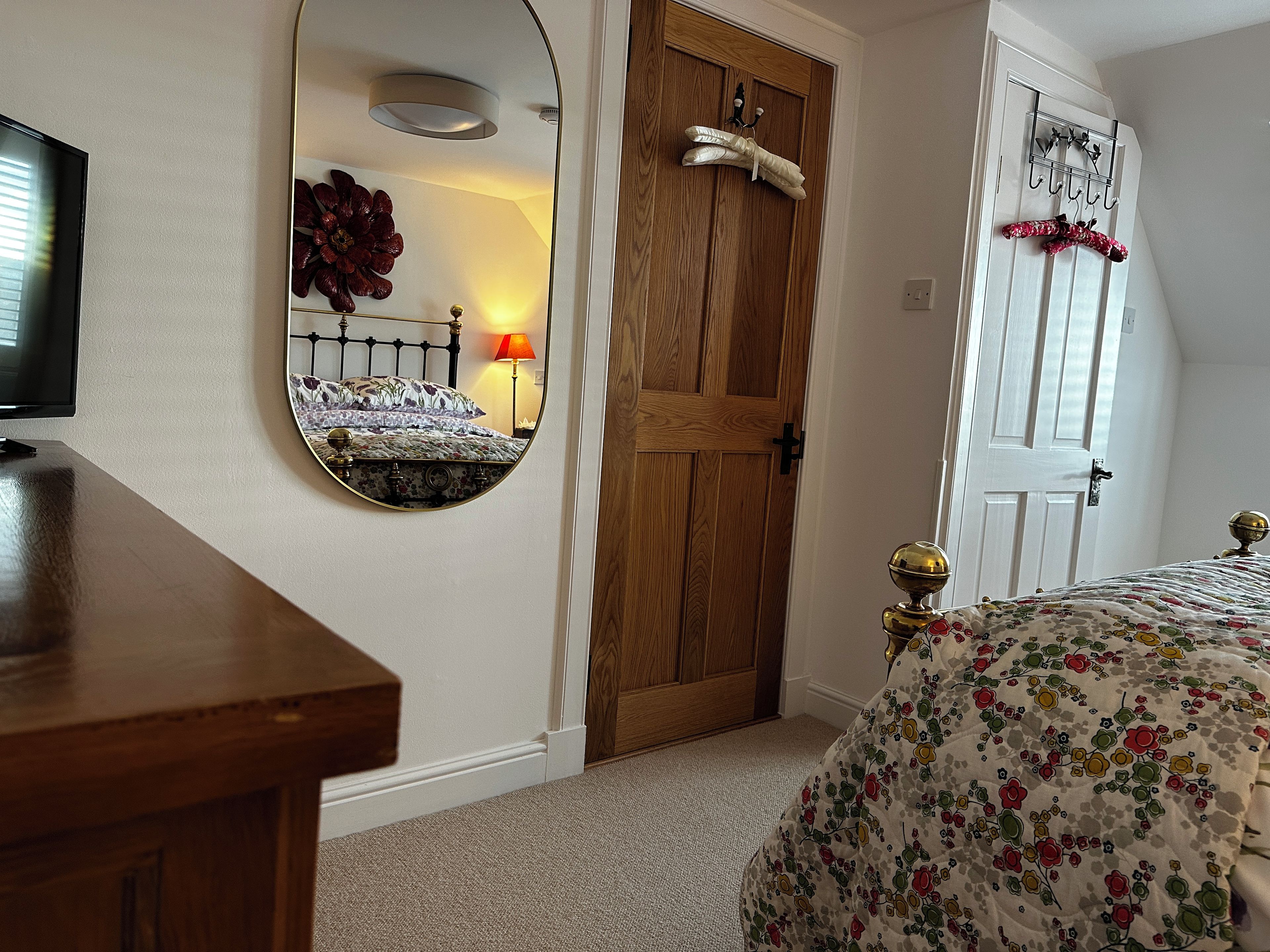 A bedroom with a wooden door, a round mirror, a bed with floral bedding, and a television on a wooden stand.