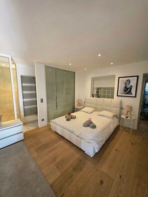 Ein modernes Schlafzimmer mit einem großen Bett, einem Glas-Kleiderschrank, einem kleinen Nachttisch und einem gerahmten Bild an der Wand.