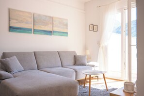 Moderni olohuone, jossa on harmaa sohva, pieni pyöreä pöytä ja suuri ikkuna, josta avautuu näkymä merelle.