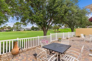 Eine Terrasse im Freien mit Tisch und Stühlen, einem weißen Zaun und Blick auf einen Golfplatz.