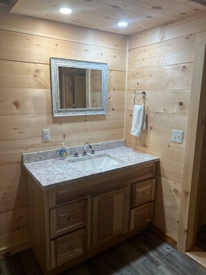 Ein Badezimmer einer Holzhütte mit Waschbecken auf einer Granitplatte, Holzschränken, einem Spiegel und einem Handtuchhalter.
