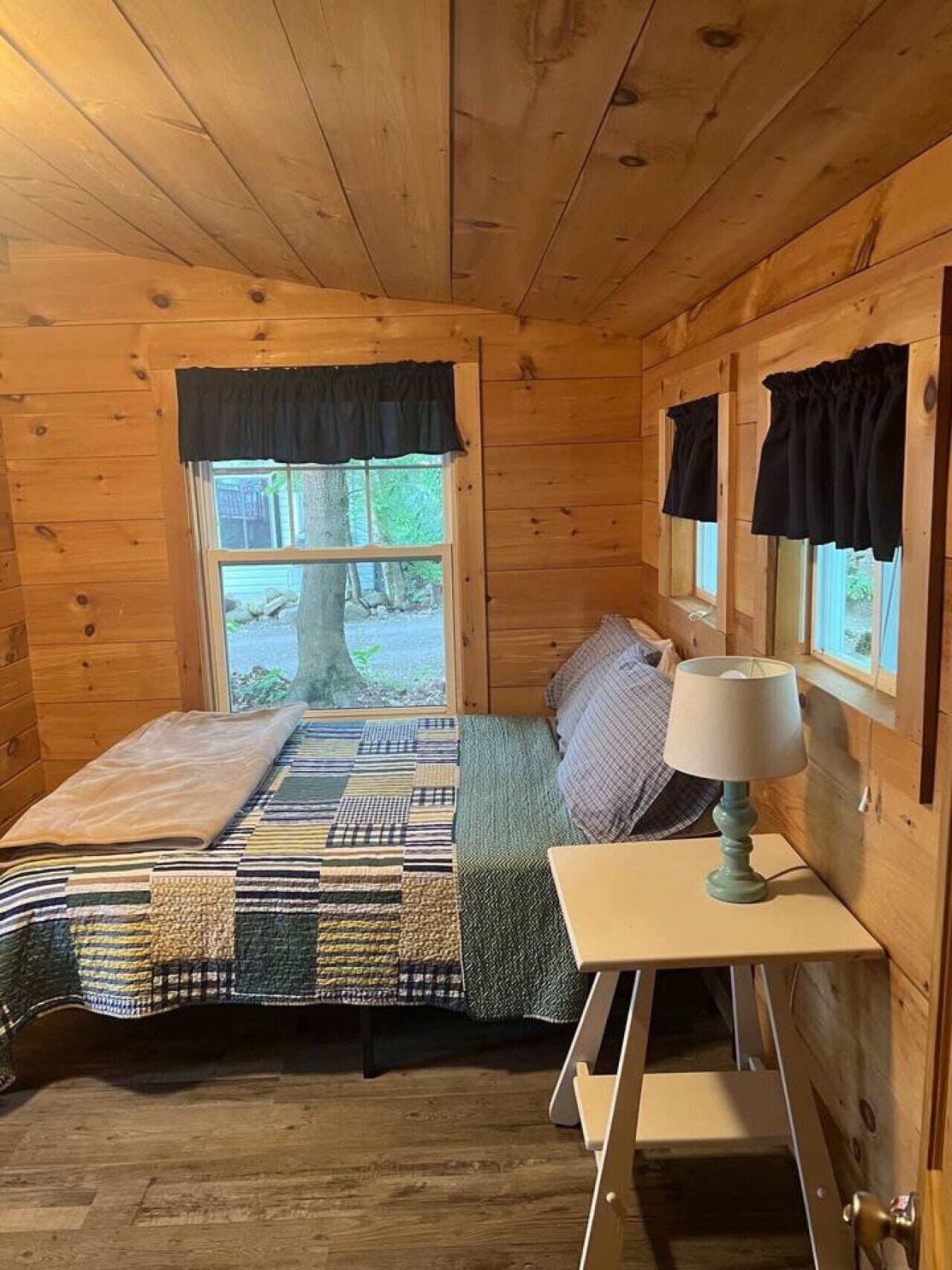 A wooden cabin bedroom with a bed, a nightstand, a lamp, and a window with a view of the outdoors.
