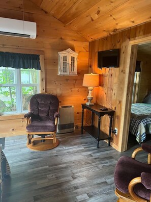Ein Zimmer in einer Holzhütte mit einem gepolsterten Sessel, einem Tisch mit einer Lampe, einem Bett und einer Klimaanlage.