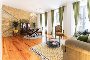 A living room with a stone wall, a round coffee table, a sofa, a chair, a dining table with chairs, and a chandelier. - Azzulea Avenida21 Historic Charm, Modern Luxury (Lisbon)