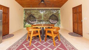A dining area with a wooden table and chairs, a ceiling fan, and a view of trees through large windows. - Walk to beach, 2BR/2BA, Apartment, pool, parking, outdoor kitchen (Playa Grande)