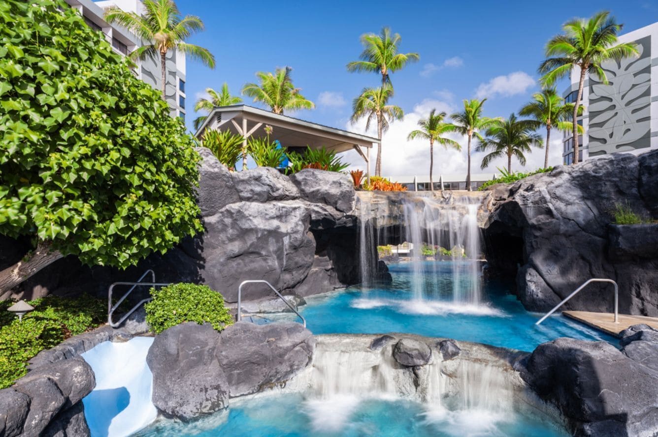 Ein Hotel mit Poolanlage, Wasserfall, Palmen und überdachter Sitzgelegenheit.