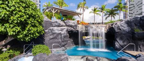 Ein Hotel mit Poolanlage, Wasserfall, Palmen und überdachter Sitzgelegenheit.