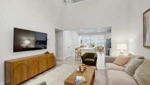 Uma sala de estar moderna com televisão de tela plana, um console de madeira, uma poltrona verde, um sofá bege e uma área de jantar ao fundo.